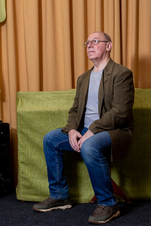 A middle-aged man in a green jacket sits on a chair near an orange curtain. Vertical photo.の写真素材