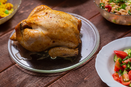 Grilled whole chicken on a glass plate on a wooden table with other food. Horizontal photo.