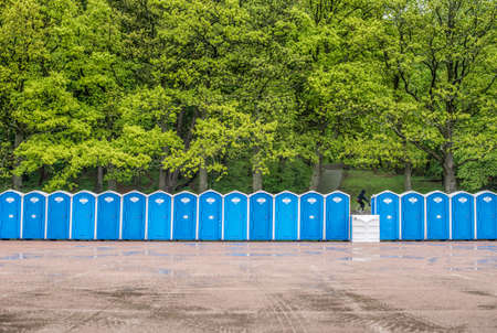 Long row of portable toilets in front of a forest where one has tipped over.の写真素材