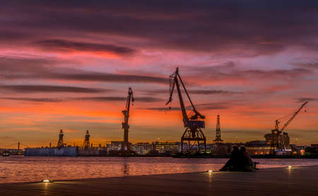 Two persons sitting dockside. In the background an old wharf with cranes