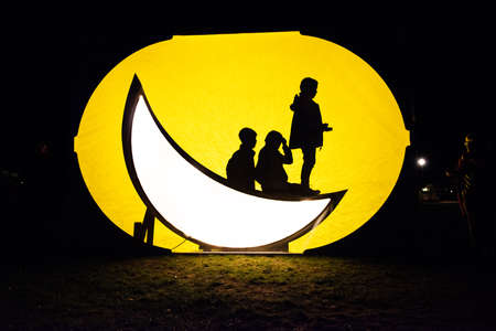 Silhouette of three young kids playing on a light sculpture formed as a moon crescent..