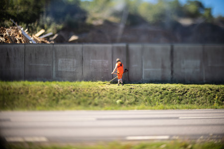Worker in high visibilty orange work wear cutting grass by a fence