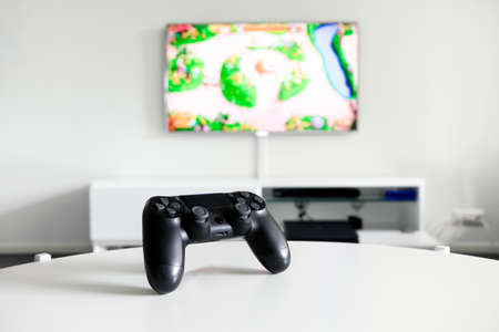 A view of a logo free, black videogame controller on a white table. Tv is in the background.