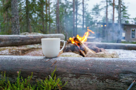 A lonely campfire burns in the cold winter forest with cottage in background. A cup of warm coffee on a log. Camping concept.の写真素材