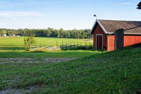 Traditional Finnish red wooden barn with roundpole fence and the weather vane on the roof. Old idyllic farm in the countryside.の写真素材