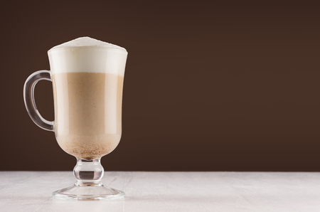 Cappuccino coffee in elegant glass with foam on white table and dark brown wall, copy space.の写真素材