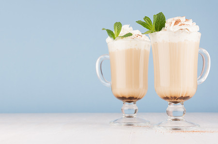 Two coffee drinks with whipped cream, green mint, cocoa powder for breakfast in glass with handle in  pastel blue interior on white wood table, copy space.の写真素材
