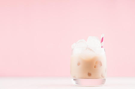 Creamy alcohol cocktail with ice cubes, striped straw in elegant shot glass on white table and soft...