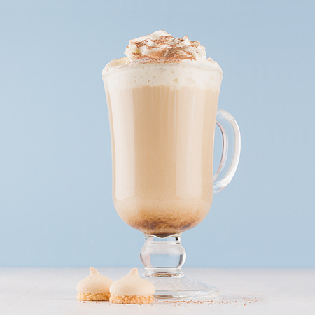 Macchiato coffee with whipped cream, chocolate powder, biscuits in transparent goblet on pastel blue background and white wooden table, square.の写真素材