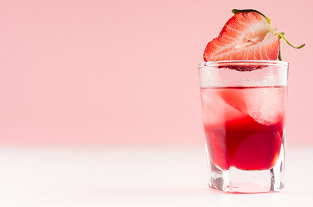 Exquisite cold red alcoholic liquor with ice cubes and strawberry slice in shot glass closeup on white wood table and pastel pink wall, copy space.の写真素材