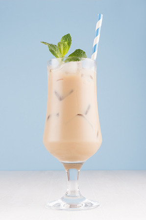 Summer cold coffee cocktail with cream, ice cubes, striped straw, green mint in transparent glass in soft pastel blue interior on white wood table, vertical.の写真素材