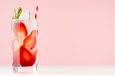 Homemade strawberry lemonade with slices, ice cubes, mint and straw in wet transparent glass in elegant soft light pink interior.