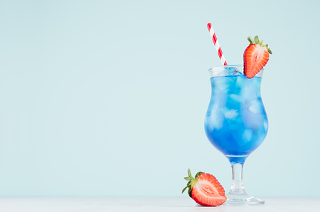 Bright blue tropical cocktail with curacao liquor, ice cubes, strawberry, straw in misted wineglass on soft light blue background.の写真素材