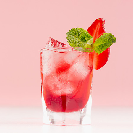 Tropical refreshing alcoholic red drink in shot glass with ice cubes, strawberry slice, green mint on white wood board and pastel pink background, square.の写真素材