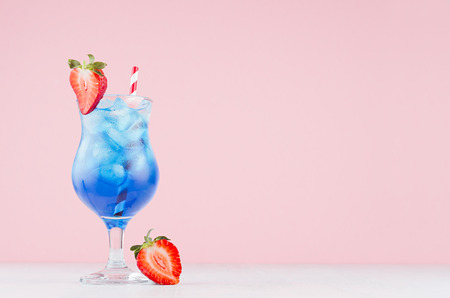 Tropical beach alcohol blue drink  - curacao liquor, strawberry slice, ice, straw in misted glass on pink background, white wood board.の写真素材