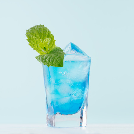 Exotic alcohol cold blue cocktail with curacao liquor, ice and green mint in rich wet shot glass on soft light pastel green background.の写真素材