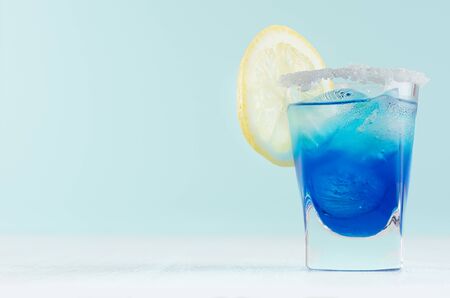 Blue curacao cocktail with lemon slice, ice cubes, sugar rim in shot glass on elegant white wood board and pastel mint color wall.の写真素材