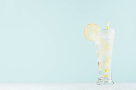 Fresh summer citrus cold drink with yellow lemon pieces, ice cubes, tonic, striped straw in light soft pastel  blue color background on white wood board.の写真素材