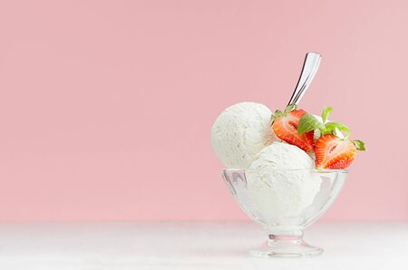 Homemade creamy ice cream scoops in glass bowl with green mint, strawberry slices, spoon in modern pink color interior on white wood board.の写真素材