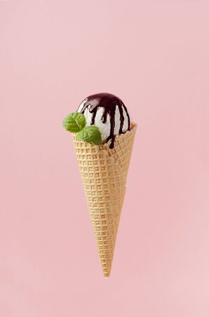 White creamy ice cream in crisp waffle cone with chocolate sauce, fresh green mint leaf on pastel pink background.