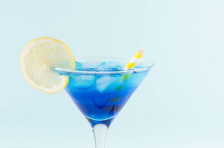 Cool summer blue beverage margarita for party with ice cubes,  lemon slice, yellow straw on white wood board and mint color wall, top part, details.の写真素材