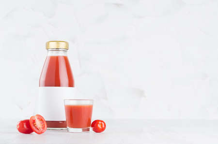 Fresh red tomato juice in glass bottle with blank label mock up with glass, vegetables slices in soft light white interior on wood table with copy space.の写真素材