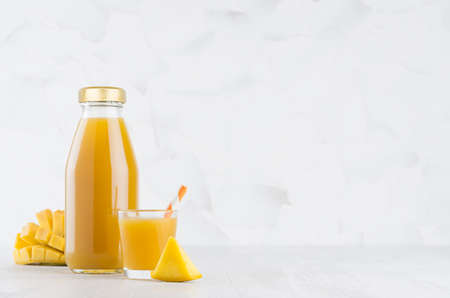 Fresh yellow mango juice in glass bottle mock up with straw, wine glass, fruit pieces on white wood table in light interior, template for packaging, advertising, design, branding product.の写真素材