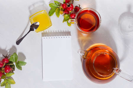 Autumn teatime with rosehip tea with teapot, cup, autumn rose hip branch and blank notepad in sunny day with shadows on white wood table top view, copy space.の写真素材