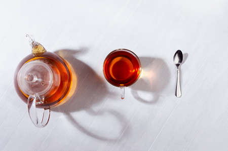 Cozy autumn atmosphere of morning teatime with black tea in transparent teapot with cup in orange sun beams and shadows on white wood table top view.の写真素材