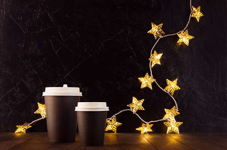 Blank black coffee paper cups with New Year gold glowing lights with stars on black plaster wall,...