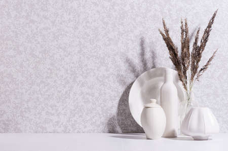 Cozy home decoration with white ceramic vase, pot, dish and bouquet of gray dried herb in morning...