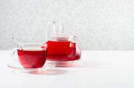 Red herbal tea in transparent tea pot with cup, spoon in sun beam in soft light white interior, copy space.の写真素材