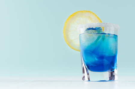Exotic cold shot glass cocktail with yellow lemon slice, blue curacao, ice cubes, salt rim on soft light mint color background and white wood board.の写真素材