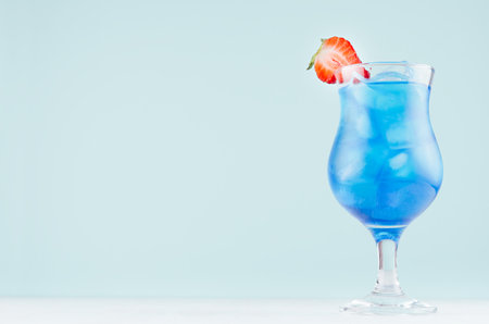 Summer blue alcohol drink with curacao liqueur, ice cubes, strawberry slice in glamor glass on soft light green background.の写真素材