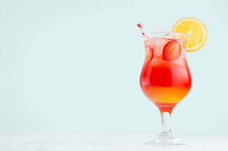 Tropical sunrise striped red, yellow drink with orange, strawberry piece, ice, straw in misted glass on pastel green wall, white wood table.の写真素材