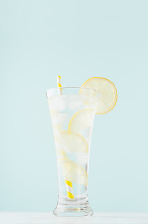 Detox cold tonic water with sunny lemon slices, ice cubes, yellow striped straw in elegant glass on white wood table, mint color wall, vertical.の写真素材