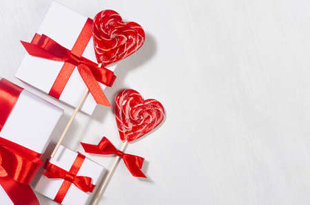 Festive love background - white gift boxes with red silk ribbon and sweet lollipops hearts on white wood board, top view, border.の写真素材