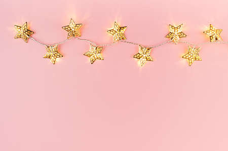 Christmas lights with golden stars glowing on soft light pastel pink background, border, copy space, top view.の写真素材