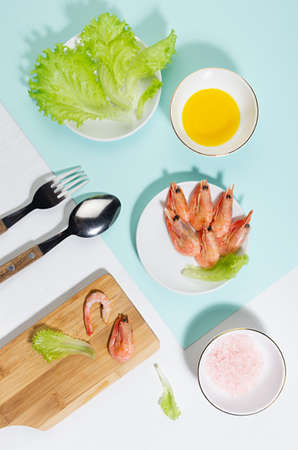 Spring fresh salad of shrimps, greens, oil, pink salt - ingredients in bowl on cutting board on mint color with shadow in sunlight. Colorful modern geometric style of food background, vertical.の写真素材