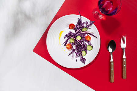 Fresh summer vegetable salad with red cabbage, cherry tomato, cucumber, oil, drink in glass with shadow in sunny day on red color, white wood table, top view.の写真素材