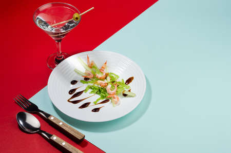 Modern elegant geometric style in food - appetizer of shrimps, greens, celery, teriyaki sauce with martini cocktail with shadow in sunlight on red and minty background, border.の写真素材