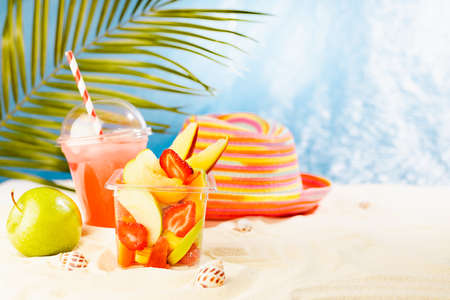 Fresh fruit salad in transparent takeaway box with cold cocktail, green apple on tropical summer beach with palm, sand, sunny sea view. Food and vacation on ocean beach.の写真素材