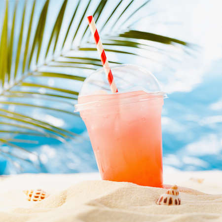 Cold tropical fruit cocktail in transparent plastic glass takeaway with straw on sunny coast with palm, sand, ocean view, square. Bright hot summer beach holiday.の写真素材