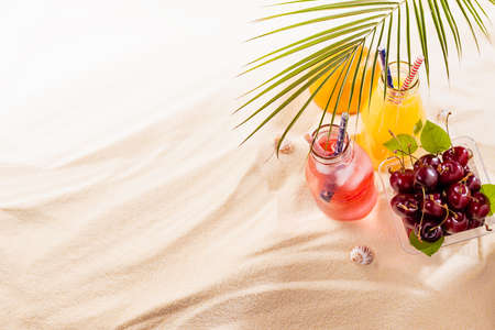 Picnic with food and drink on tropical beach, vacation background - fresh cold drinks with straw in glass bottles, green palm leaf and ripe red cherry, orange in box on white sand, copy space.の写真素材
