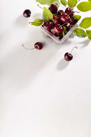 Summer takeaway food - wet ripe red fresh cherry in plastic box with green leaves on white wood...