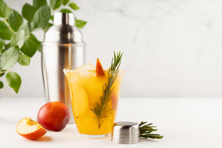 Bright fruit summer beverage mixed with ripe peach, ice, green rosemary twig, sugar rim, ingredients, silver shaker, green foliage in sunlight in modern soft light white bar interior or outdoor.の写真素材