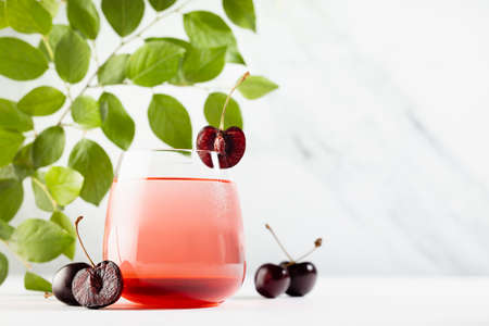 Fresh summer cold cherry cocktail with green foliage, slice fruit in golden sunshine in elegant white kitchen interior with soft light marble wall or outdoor.の写真素材