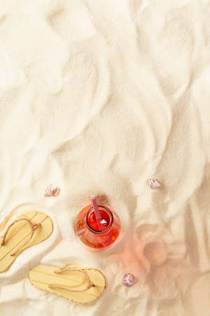 Tropical picnic and recreation on sandy beach - fresh cold fruit pink cocktail in glass bottle, slippers, seashells on sand, border, top view, vertical.の写真素材