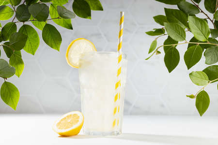 Fresh summer homemade lemonade in misted glass with ice, straw with green leaves in bright sunlight on white kitchen or outdoor.の写真素材