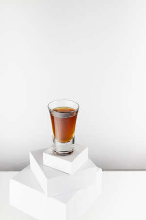Bright brown brandy in glossy shot glass in sunlight with reflections on abstract white square...